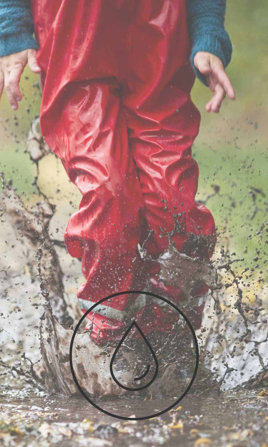 Child wearing green waterproof rain gear and mittens splashing in shallow water at the shoreline.