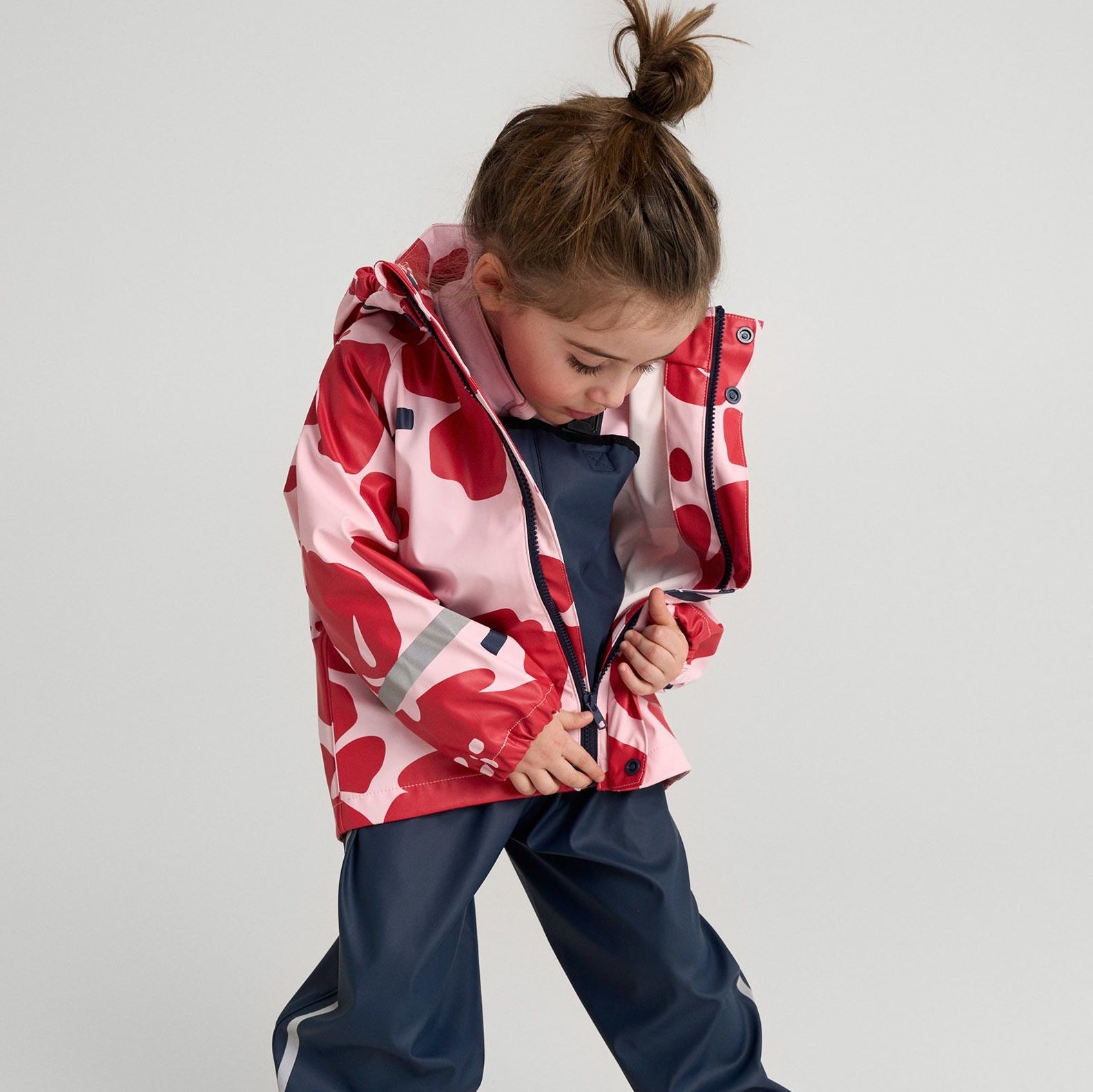 Smiling young girl wearing a pink Helly Hansen rain jacket with hood up, standing outdoors on a rainy day.