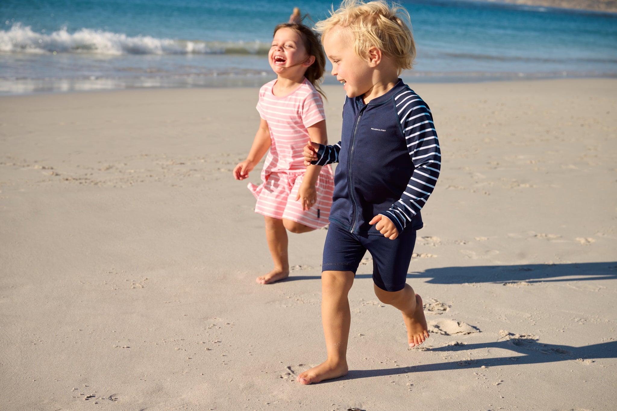 Two children running and laughing on a sunny beach wearing protective swimwear
