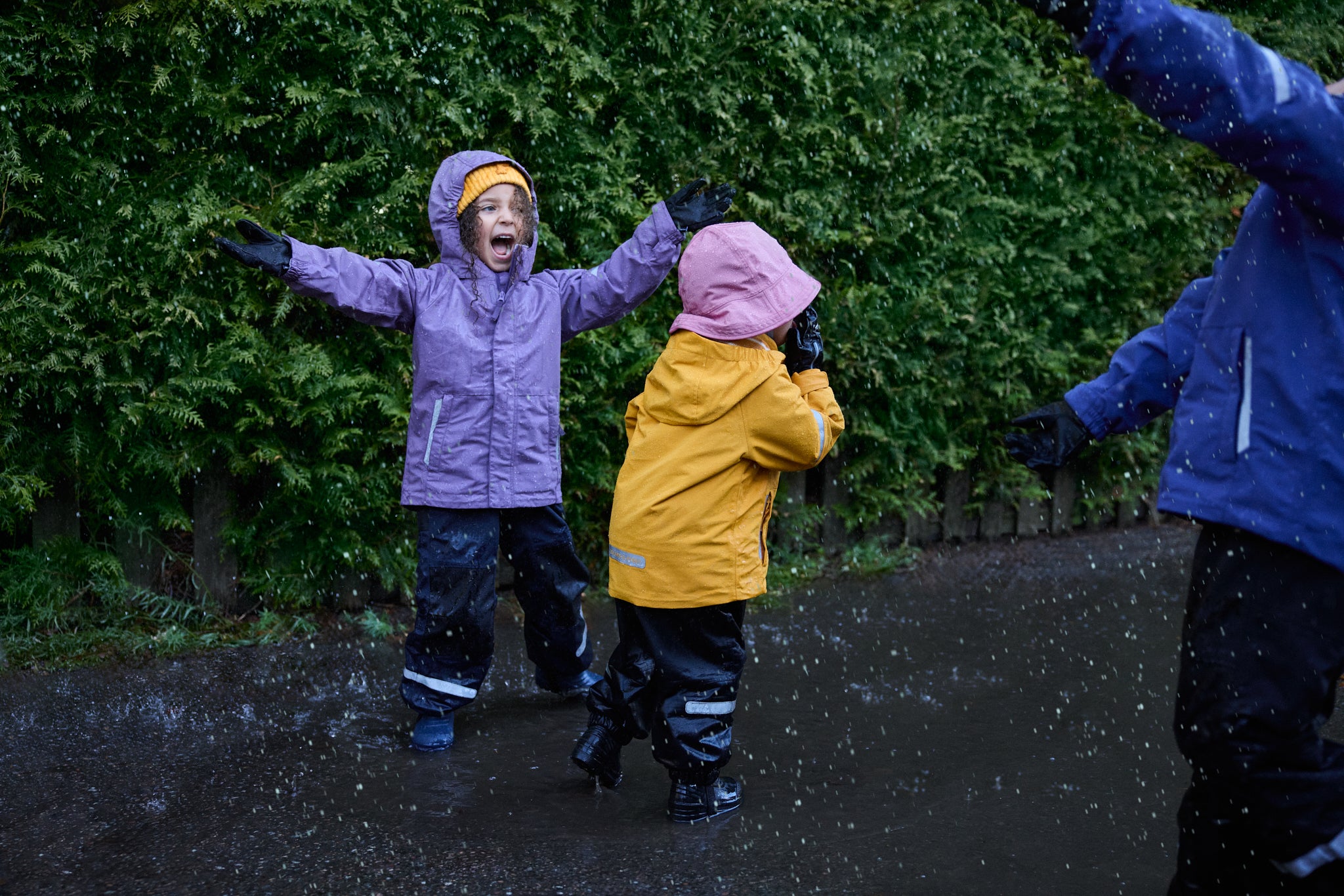 Children playing in the rain with colorful jackets against a green hedge.