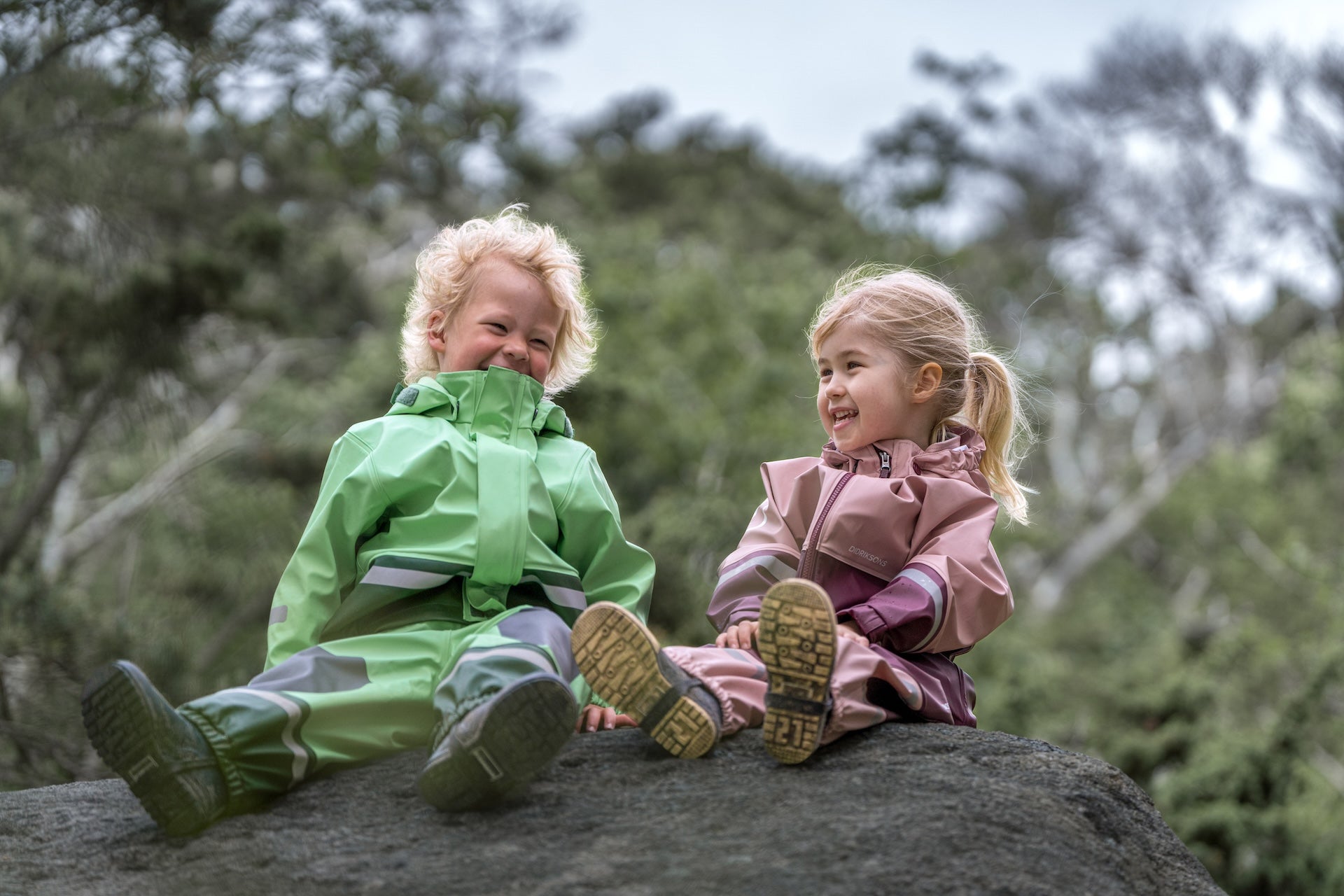 Two children in colorful raincoats sitting on a rock with trees in the background