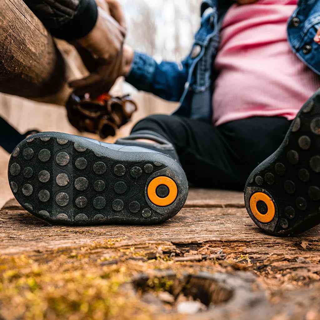 Close-up of a child’s black rain boots with circular orange tread, sitting outdoors on a wooden surface next to an adult holding their hand.