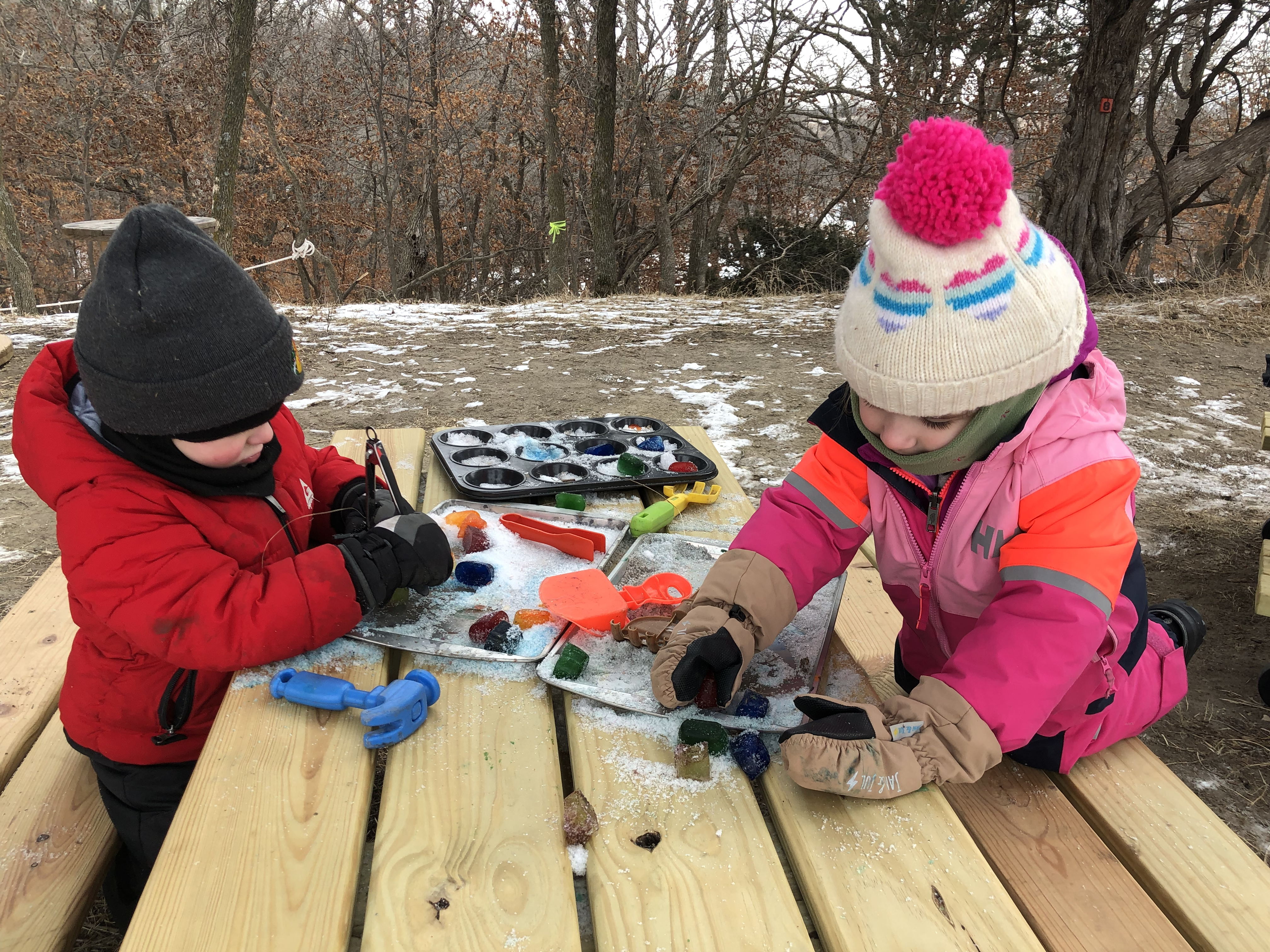 Two children playing with art supplies on a wooden table outdoors.