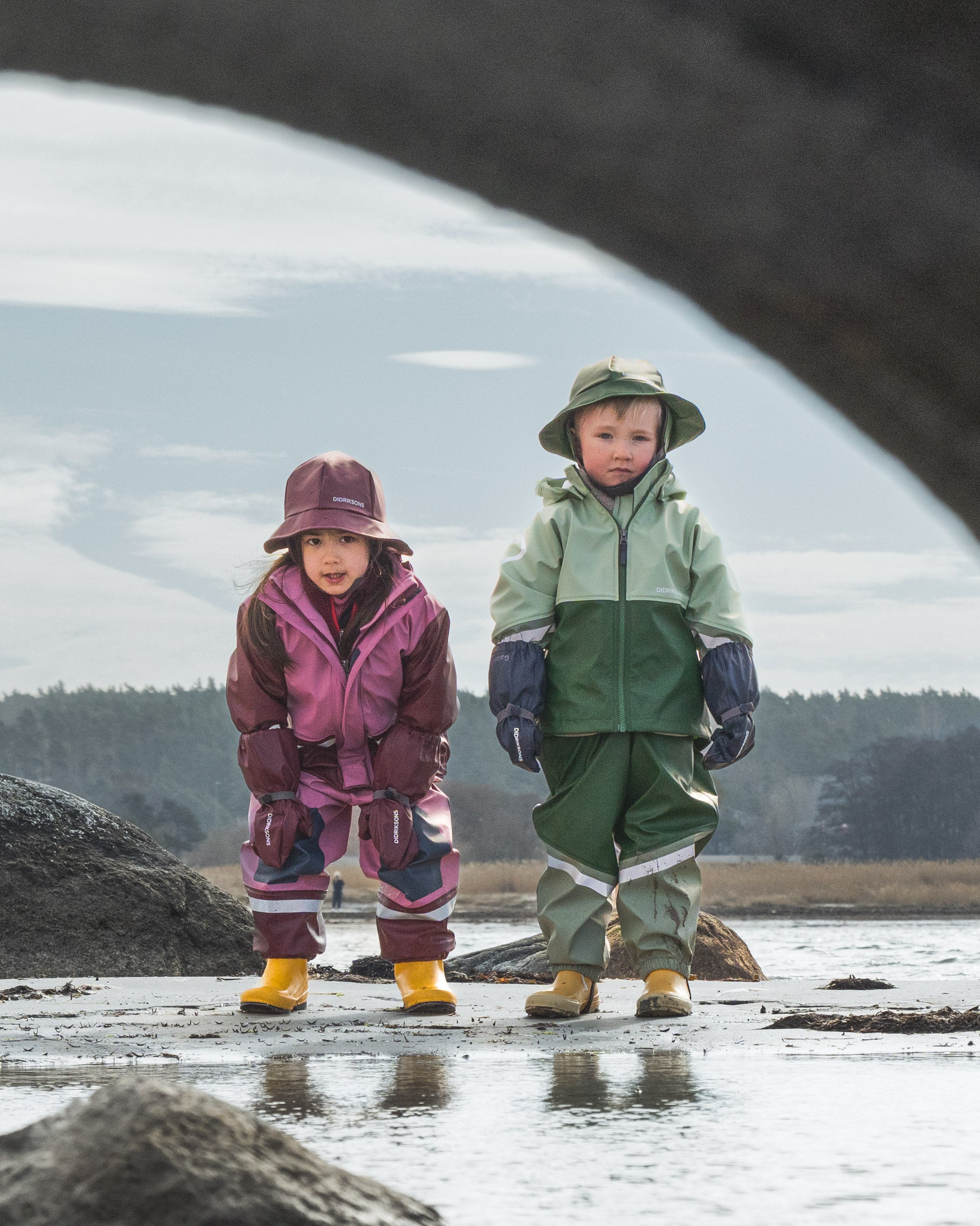 Two children in waterproof suits standing on a wetland with a bridge in the background.